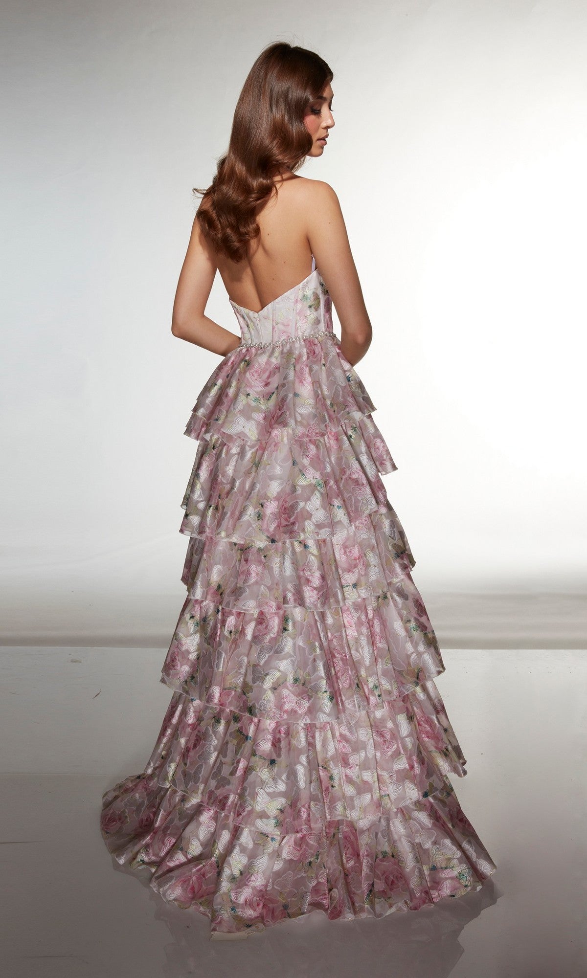 A woman stands with her back to the camera, wearing the Alyce 61996 long prom dress, a strapless, floor-length gown with tiered pink and white floral organza.
